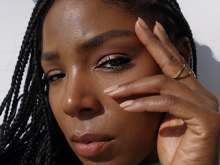 Picture of a person with dark skin wearing nude nail polish, holding their fingers to their face