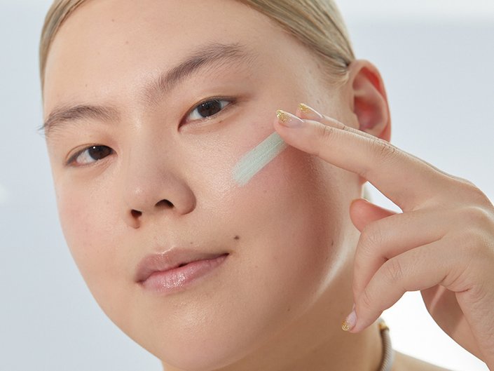 Person with blonde hair pulled back, looking at the camera and swatching green color-correcting concealer on their cheek