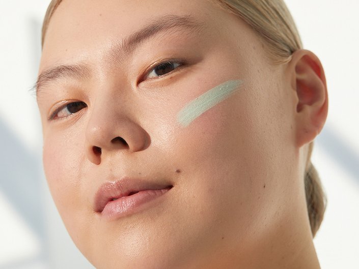 Person with blonde hair pulled back and green color-correcting makeup swatched on their cheek