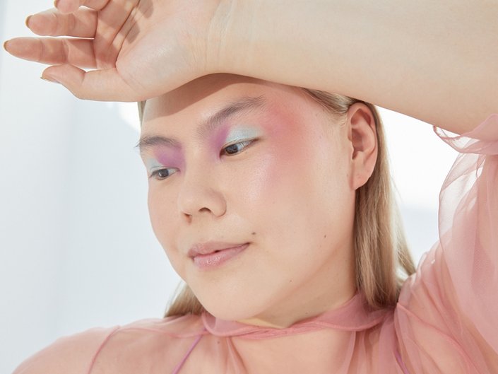 Picture of a model with mint green and lavender eyeshadow and pink blush looking off to the side with a hand held to their forehead